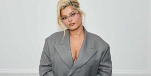 Bebe Rexha cantando en un escenario iluminado con pantallas de fondo y vestuario brillante.